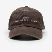 A brown denim cap with the text 'DOGS. BECAUSE PEOPLE SUCK' embroidered on the front.